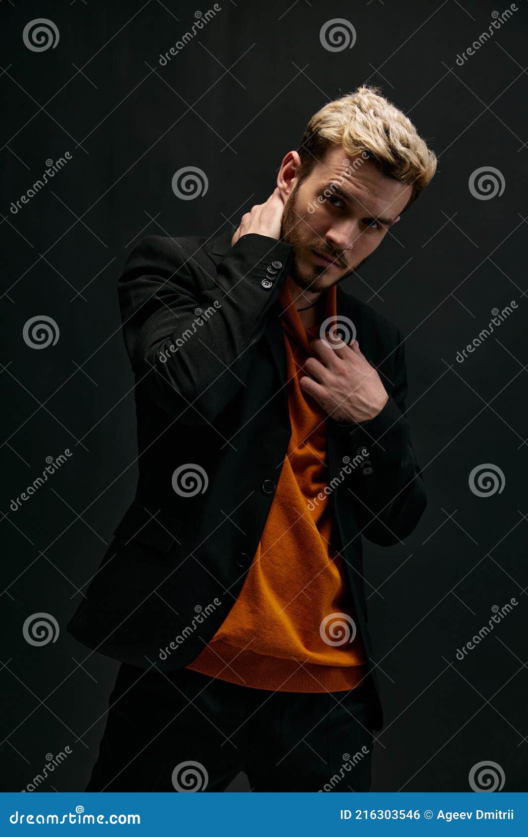 1067x1690  Блондинный парень в апельсиновом свитере и куртке на темном фоне держит руку за его головой stock photo - изображение света, портрет: 216303546