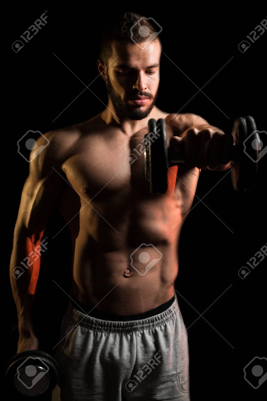 867x1300  Молодой человек тренируется плечами с гантелями на черном фоне. Изображение 81239211.