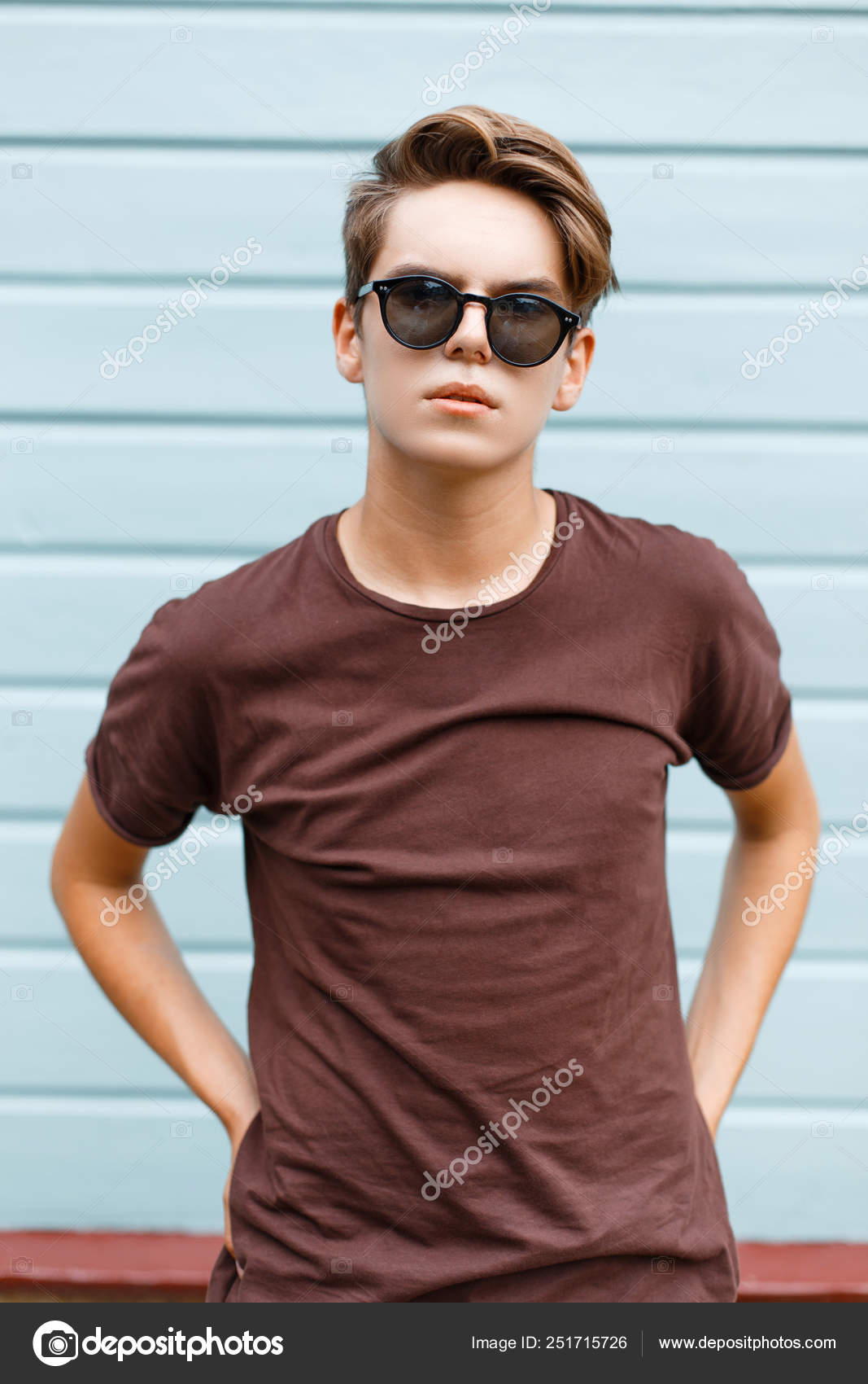 1067x1700  Модный современный молодой хипстер в темных солнцезащитных очках в модной  летней футболке со стильной прической стоит рядом со старинной голубой  деревянной стеной. Красивый парень на улице. Летний стиль . стоковое фото  ©alonesdj